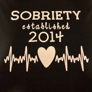 May include: Black t-shirt with white text that reads "SOBRIETY established 2014" and a white heart with a heartbeat graphic.