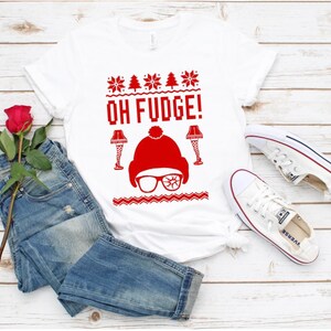 Może przedstawiać: Biała koszulka z czerwonym wzorem świątecznego swetra z napisem "OH FUDGE!" i kreskówkowym obrazkiem osoby w czapce Świętego Mikołaja i okularach. Koszulka leży na białym drewnianym tle z parą białych butów sportowych i czerwoną różą.