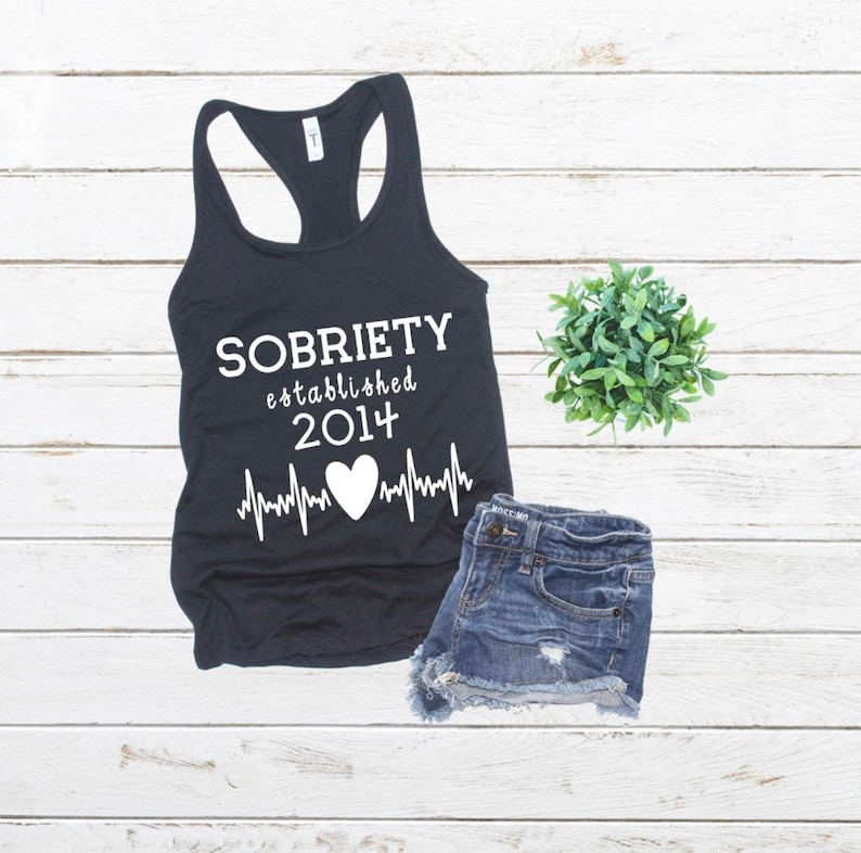 May include: Black tank top with white text that reads "SOBRIETY established 2014" with a heart and a heartbeat graphic.