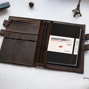 以下が含まれることがあります： 茶色の革製ジャーナルカバー。中には黒色の2025年ウィークリープランナーが入っています。プランナーには「LEUCHTTURM1917」と「Weekly Planner」の文字が入った白いラベルが付いています。カバーにはポケットとジッパーが付いています。
