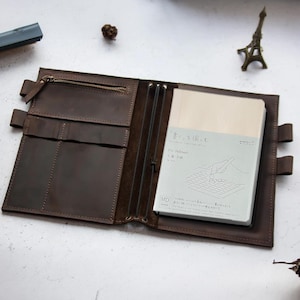 May include: A brown leather notebook cover with a zippered pocket, multiple elastic loops, and a pen holder. The cover is open to reveal a blank notebook with the text "書くことは楽しい" and "MD Notebook" printed on the front.