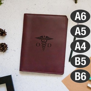 May include: A burgundy leather journal with a gold embossed medical symbol and the letters "OD". The journal is on a white surface with pine cones and a brown envelope.
