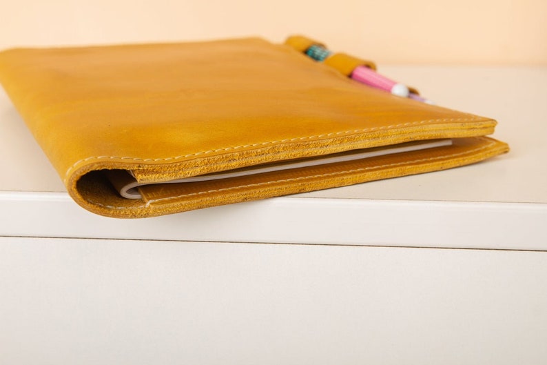 May include: A yellow leather bound notebook with a pen holder and two pens sticking out.