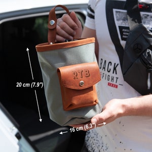 Op de afbeelding: Een grijze canvas tas met een bruine leren flap en zak. De zak heeft de initialen "RTB" erop gegraveerd. De tas is 20 cm hoog en 16 cm breed.