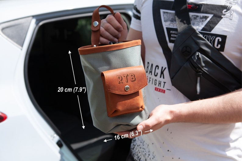 Op de afbeelding: Een grijze canvas tas met een bruine leren flap en zak. De zak heeft de initialen "RTB" erop gegraveerd. De tas is 20 cm hoog en 16 cm breed.