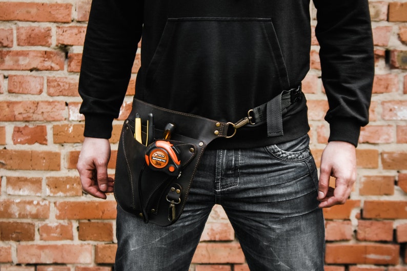 Pode incluir: Um cinto de ferramentas de couro castanho com uma variedade de bolsos e la&ccedil;os. O cinto &eacute; usado sobre uma camisa preta e cal&ccedil;as de ganga azuis. O cinto tem uma fita m&eacute;trica, uma chave de fenda e outras ferramentas presas a ele.
