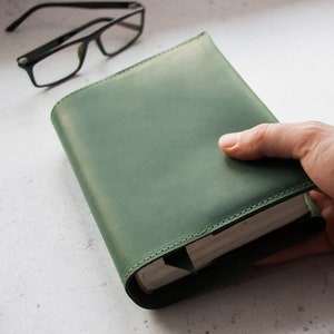 May include: A green leather-bound journal with a hand holding the book. The journal has a stitched edge and a simple design.