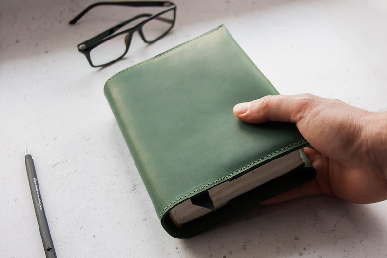 May include: A green leather-bound journal with a hand holding the book. The journal has a stitched edge and a simple design.