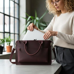 May include: A burgundy leather laptop bag with a shoulder strap and top handles. The bag features a front pocket and a zip closure. A silver laptop is being placed inside the bag.
