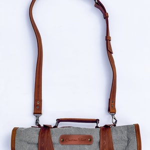 May include: A gray canvas roll-up bag with a brown leather strap and handle. The bag has a brown leather patch with the text "Custom Text" on it.