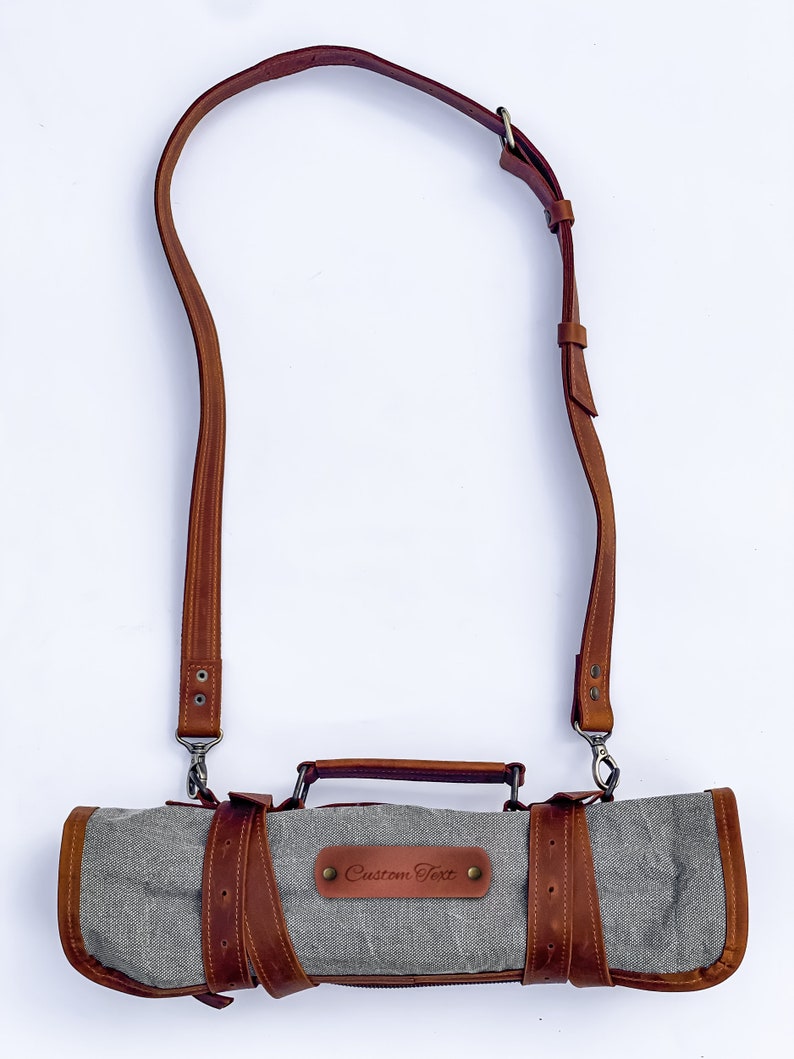 May include: A gray canvas roll-up bag with a brown leather strap and handle. The bag has a brown leather patch with the text "Custom Text" on it.