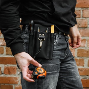 K&ouml;nnte beinhalten: Ein schwarzer Leder-Werkzeugg&uuml;rtel mit mehreren Taschen und einem Metallclip. Der G&uuml;rtel wird &uuml;ber einer blauen Jeans getragen. Ein kleines orangefarbenes Ma&szlig;band wird in der Hand gehalten.