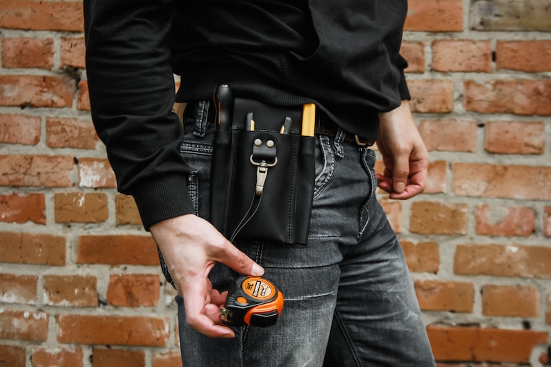 K&ouml;nnte beinhalten: Ein schwarzer Leder-Werkzeugg&uuml;rtel mit mehreren Taschen und einem Metallclip. Der G&uuml;rtel wird &uuml;ber einer blauen Jeans getragen. Ein kleines orangefarbenes Ma&szlig;band wird in der Hand gehalten.