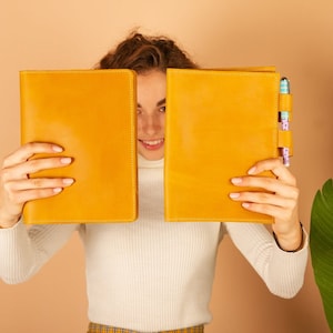 May include: A person is holding two yellow leather-bound journals in front of their face. The journal on the right has a pen holder with two pens.