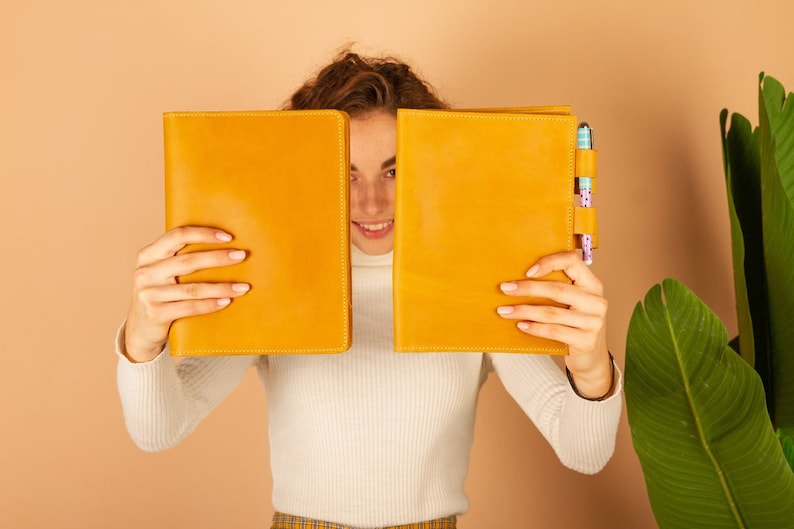 May include: A person is holding two yellow leather-bound journals in front of their face. The journal on the right has a pen holder with two pens.
