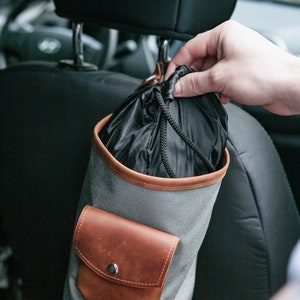 Op de afbeelding: Een grijze canvas tas met een bruine leren klep en een trekkoord. De tas hangt aan de hoofdsteun van een autostoel.