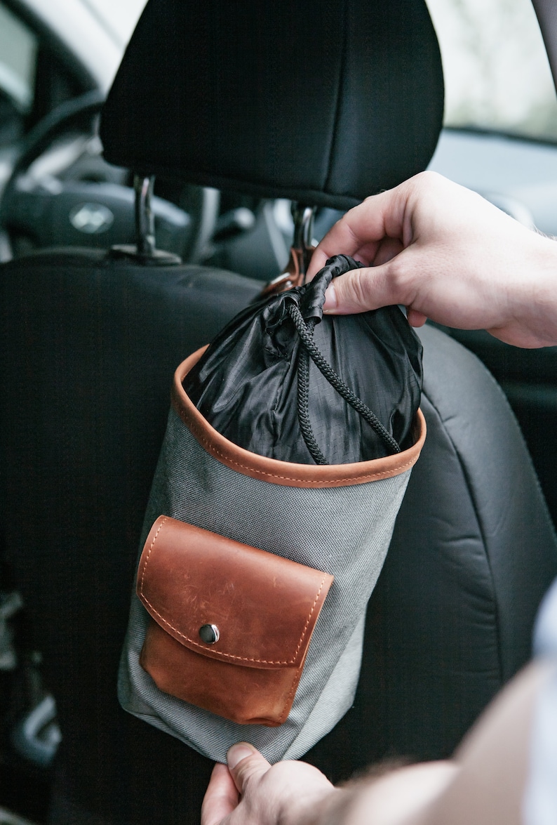 Op de afbeelding: Een grijze canvas tas met een bruine leren klep en een trekkoord. De tas hangt aan de hoofdsteun van een autostoel.