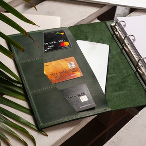Pode incluir: Um fich&aacute;rio de couro verde escuro com um mecanismo de argolas, tr&ecirc;s cart&otilde;es de cr&eacute;dito, um smartphone e um bloco de notas. O fich&aacute;rio tem v&aacute;rios bolsos para guardar cart&otilde;es e outros itens.