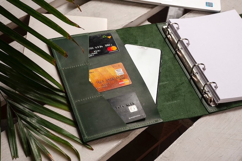 Pode incluir: Um fich&aacute;rio de couro verde escuro com um mecanismo de argolas, tr&ecirc;s cart&otilde;es de cr&eacute;dito, um smartphone e um bloco de notas. O fich&aacute;rio tem v&aacute;rios bolsos para guardar cart&otilde;es e outros itens.