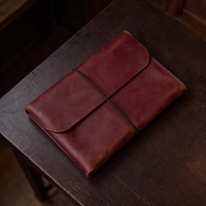 May include: A burgundy leather journal with a flap closure and a black elastic band. The journal is closed and resting on a dark wooden surface. The leather has a slightly distressed appearance.
