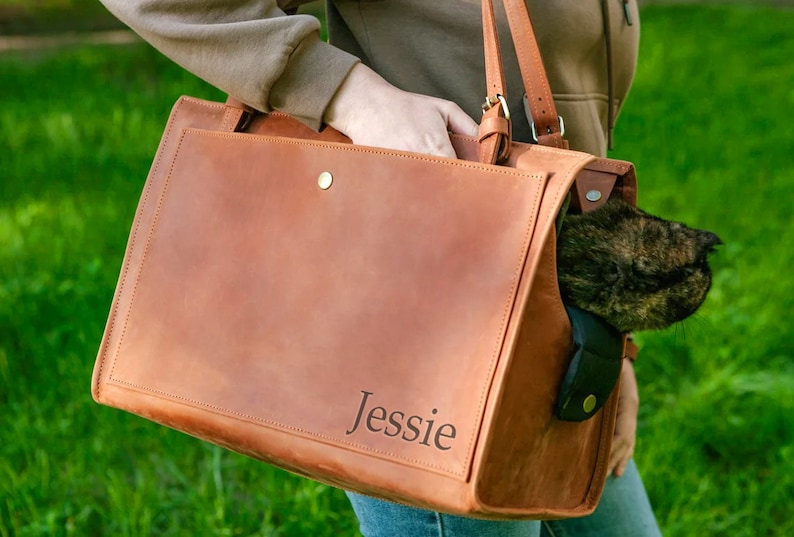 May include: A tan leather pet carrier bag with a shoulder strap and a flap closure. The bag is personalised with the name "Jessie". A cat is visible inside the bag.