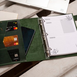 Pode incluir: Uma agenda encadernada em couro verde escuro com um anel de encaderna&ccedil;&atilde;o prateado. A agenda tem um bolso na capa frontal que cont&eacute;m cart&otilde;es de cr&eacute;dito e um telefone. A agenda est&aacute; aberta em uma p&aacute;gina em branco com papel pautado e um pequeno desenho de um dente de alho.