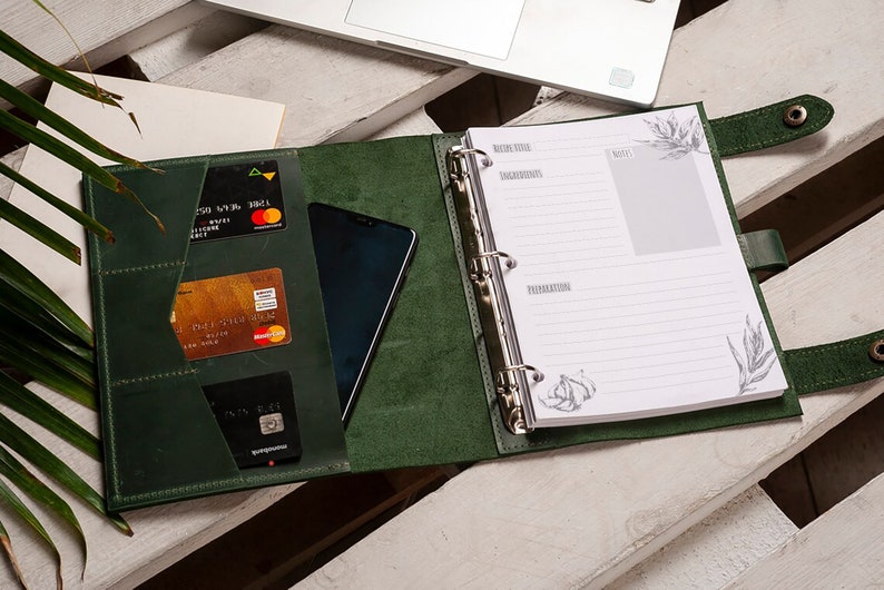 Pode incluir: Uma agenda encadernada em couro verde escuro com um anel de encaderna&ccedil;&atilde;o prateado. A agenda tem um bolso na capa frontal que cont&eacute;m cart&otilde;es de cr&eacute;dito e um telefone. A agenda est&aacute; aberta em uma p&aacute;gina em branco com papel pautado e um pequeno desenho de um dente de alho.