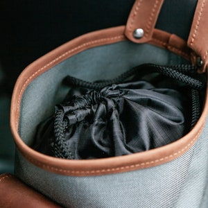 Op de afbeelding: Een grijze canvas tas met bruine leren afwerking en een zwart trekkoordzakje erin. De tas heeft een lusvormige handgreep en is ontworpen om aan de achterkant van een autostoel te hangen.