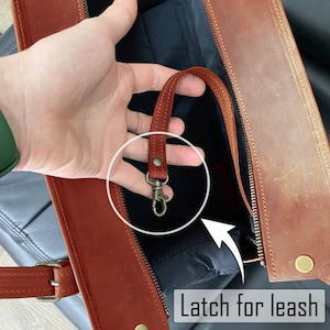 May include: Close-up of a brown leather bag with a zip and a small handle. The bag has a metal latch for a leash. The text "Latch for leash" is visible.