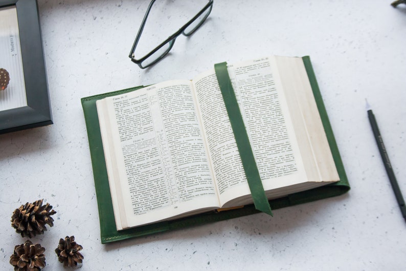 May include: A green leather-bound journal with a bookmark, open to a page with text. The journal is on a white surface with a pair of glasses, a pencil, and pine cones.