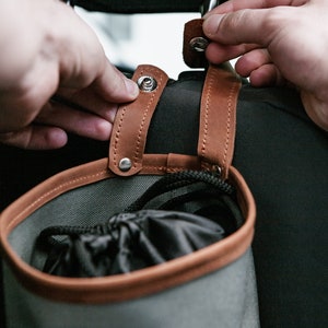 Op de afbeelding: Een grijze canvas tas met bruine leren afwerking en banden. De tas heeft een trekkoord sluiting en is bevestigd aan de achterkant van een stoel met metalen drukknopen.