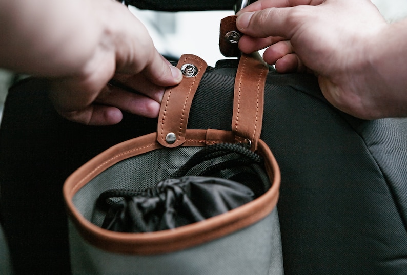 Op de afbeelding: Een grijze canvas tas met bruine leren afwerking en banden. De tas heeft een trekkoord sluiting en is bevestigd aan de achterkant van een stoel met metalen drukknopen.
