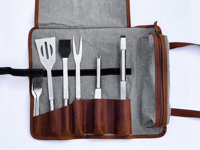 May include: A brown leather roll-up case with a gray canvas exterior and a zipper closure. The case contains a set of six grilling tools, including a spatula, a basting brush, a fork, a knife, a tongs, and a cleaning tool.