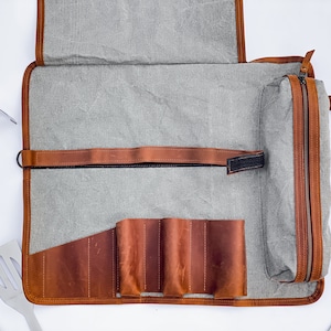 May include: A gray canvas roll-up bag with a brown leather zippered pouch and a brown leather strap. The bag has multiple slots for holding tools, such as knives or grilling utensils.