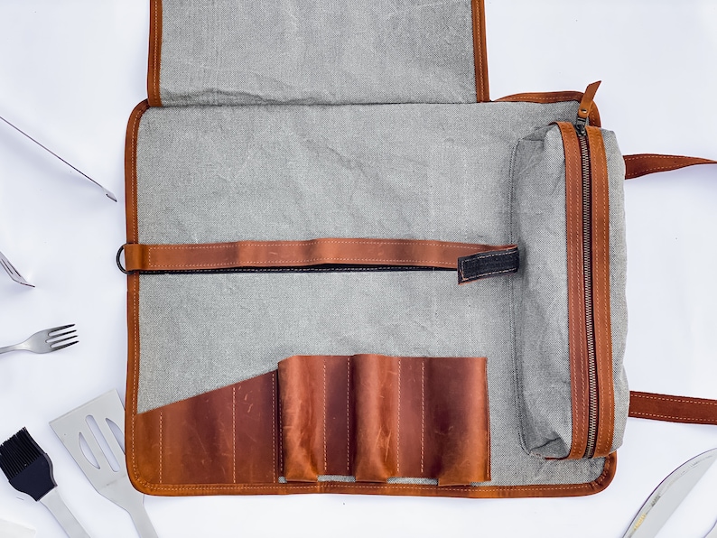 May include: A gray canvas roll-up bag with a brown leather zippered pouch and a brown leather strap. The bag has multiple slots for holding tools, such as knives or grilling utensils.