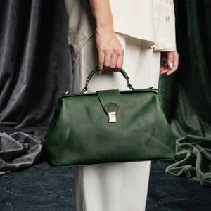 May include: A dark green leather doctor bag with a top handle and a gold-colored clasp. The bag has a rectangular shape with rounded edges. The bag is held by a person wearing a white outfit.
