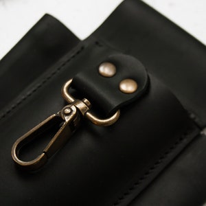 K&ouml;nnte beinhalten: Schwarze Ledertasche mit einem Messinghaken und zwei Messingnieten. Die Tasche hat eine gen&auml;hte Naht an den R&auml;ndern.