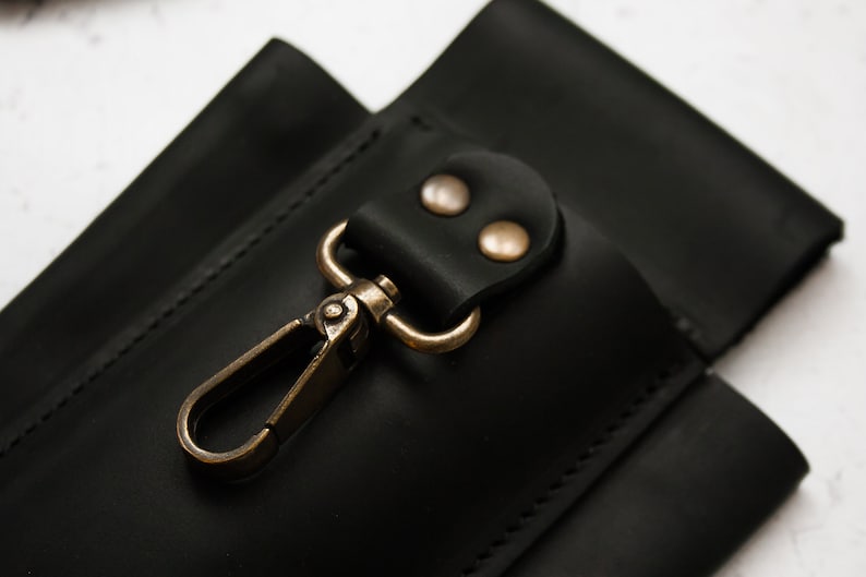K&ouml;nnte beinhalten: Schwarze Ledertasche mit einem Messinghaken und zwei Messingnieten. Die Tasche hat eine gen&auml;hte Naht an den R&auml;ndern.