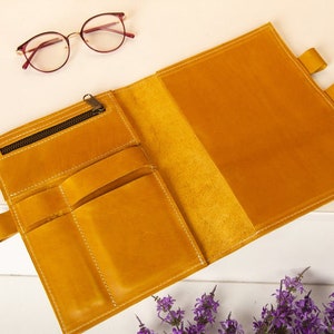 May include: A yellow leather notebook with a zippered pocket and multiple slots for pens and pencils. The notebook is open to reveal a blank page.