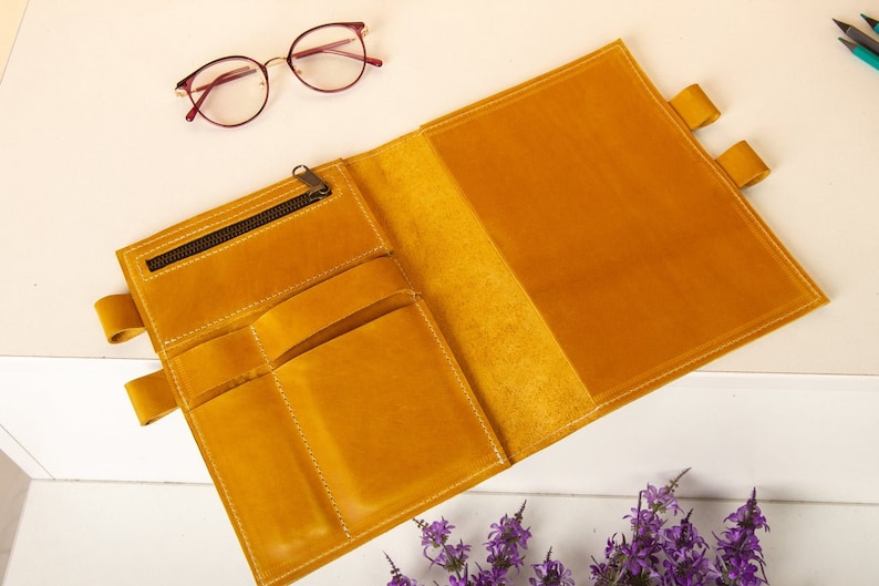 May include: A yellow leather notebook with a zippered pocket and multiple slots for pens and pencils. The notebook is open to reveal a blank page.