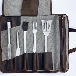 May include: A brown leather grilling tool roll with a zipper closure and a gray canvas interior. The roll contains five grilling tools: a pair of tongs, a spatula, a basting brush, a fork, and a knife.