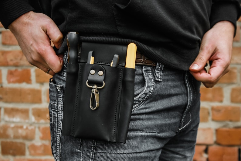 K&ouml;nnte beinhalten: Ein schwarzer Leder-Werkzeugg&uuml;rtel mit einem Metallclip und verschiedenen Werkzeugen, darunter ein gelber Schraubendreher und ein schwarzes Universalmesser. Der G&uuml;rtel wird &uuml;ber einer dunkelgrauen Jeans getragen.