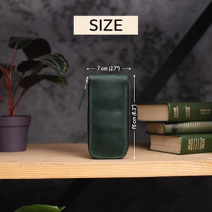 Może przedstawiać: Etui na oł&oacute;wki ze sk&oacute;ry w ciemnozielonym kolorze z zamkiem błyskawicznym. Etui ma 7 cm szerokości i 16 cm wysokości.
