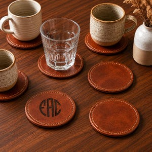 May include: A selection of brown leather coasters with white stitching. Some coasters feature a monogram design. The image also shows ceramic mugs and a glass of water, all resting on the coasters. The coasters are round and appear to be made of genuine leather.