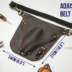 Pode incluir: Cinto de ferramentas de couro marrom com uma al&ccedil;a ajust&aacute;vel. O cinto tem um bolso grande e um bolso menor. O cinto tem 24 cm de largura e 27 cm de altura. O texto CINTUR&Atilde;O AJUST&Aacute;VEL &eacute; vis&iacute;vel na imagem.