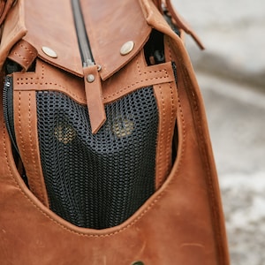 May include: Close-up of a brown leather pet carrier bag with a black mesh window. The bag has a zip closure and metal rivets. The interior is visible through the mesh.