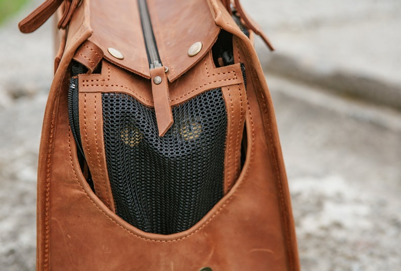 May include: Close-up of a brown leather pet carrier bag with a black mesh window. The bag has a zip closure and metal rivets. The interior is visible through the mesh.