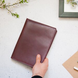 May include: A brown leather bound journal or notebook with a hand holding it open. The journal is on a white surface with pine cones and a branch with white flowers.
