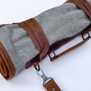 May include: A gray canvas roll-up bag with brown leather straps and a handle. The bag has a metal clasp and a shoulder strap.