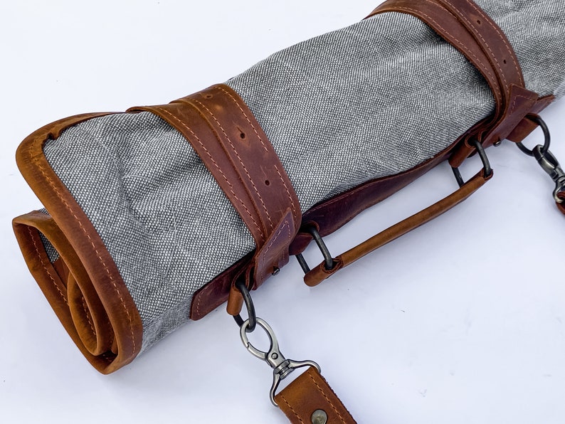 May include: A gray canvas roll-up bag with brown leather straps and a handle. The bag has a metal clasp and a shoulder strap.